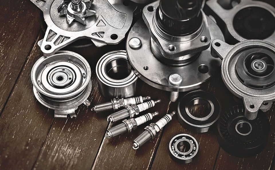 An assortment of metallic automotive parts, including spark plugs, bearings, a water pump, and a wheel hub assembly, neatly arranged on a dark wooden surface.